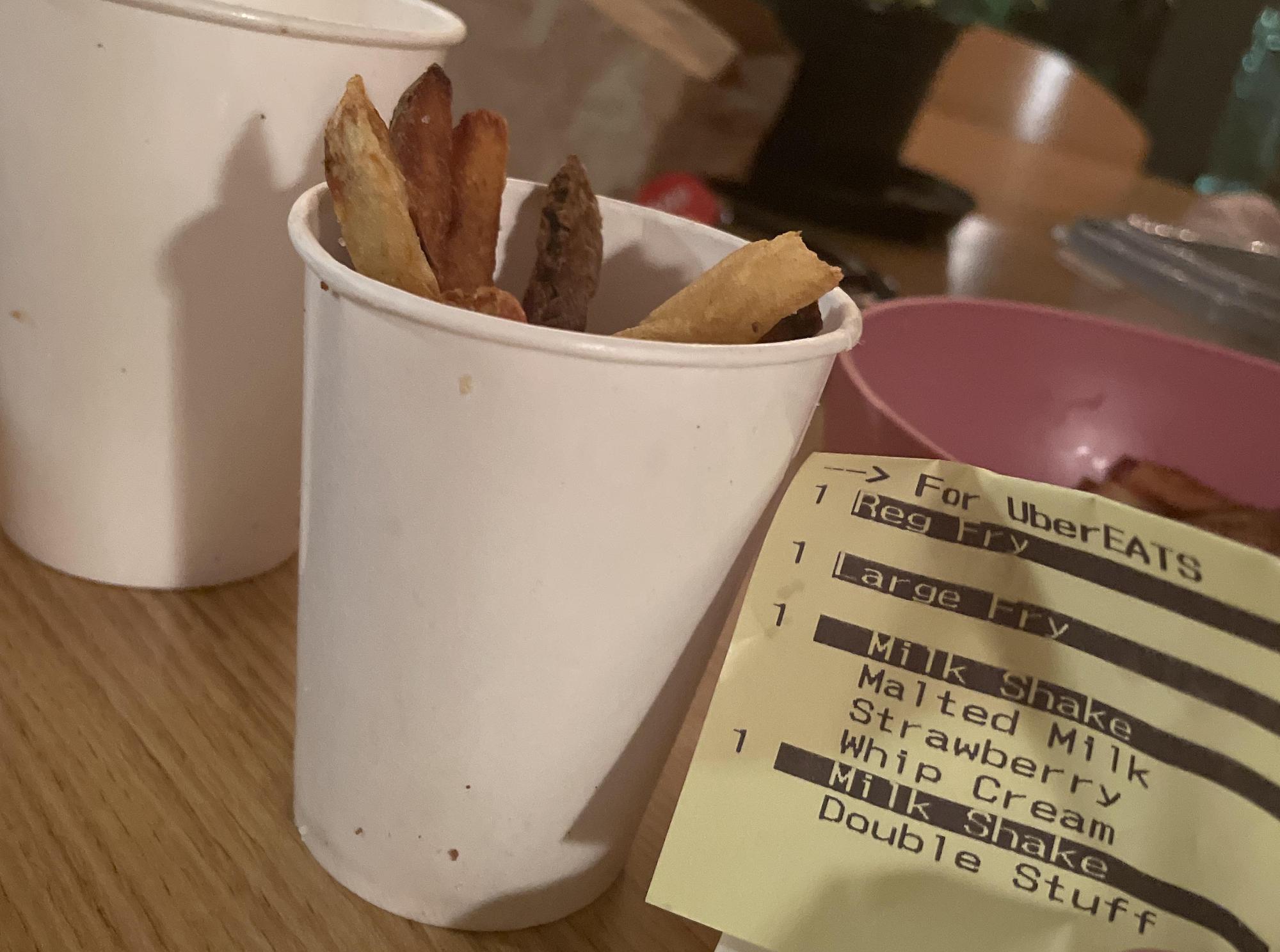 A Five Guys white cup filled with fries sits on a wooden table next to another cup and a pink bowl. A printed UberEATS receipt listing fries and milkshakes is partially visible in the foreground.