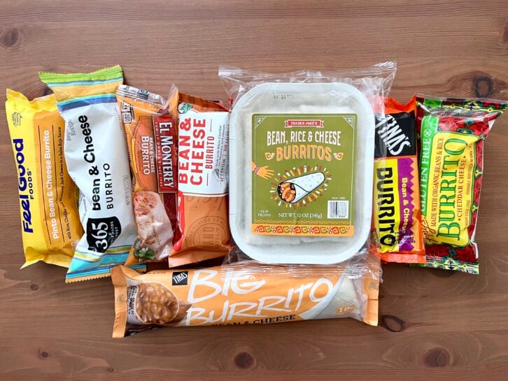 Eight packaged bean and cheese burritos from different brands are arranged on a wooden surface, displayed side by side with colorful wrappers showing their labels and branding.