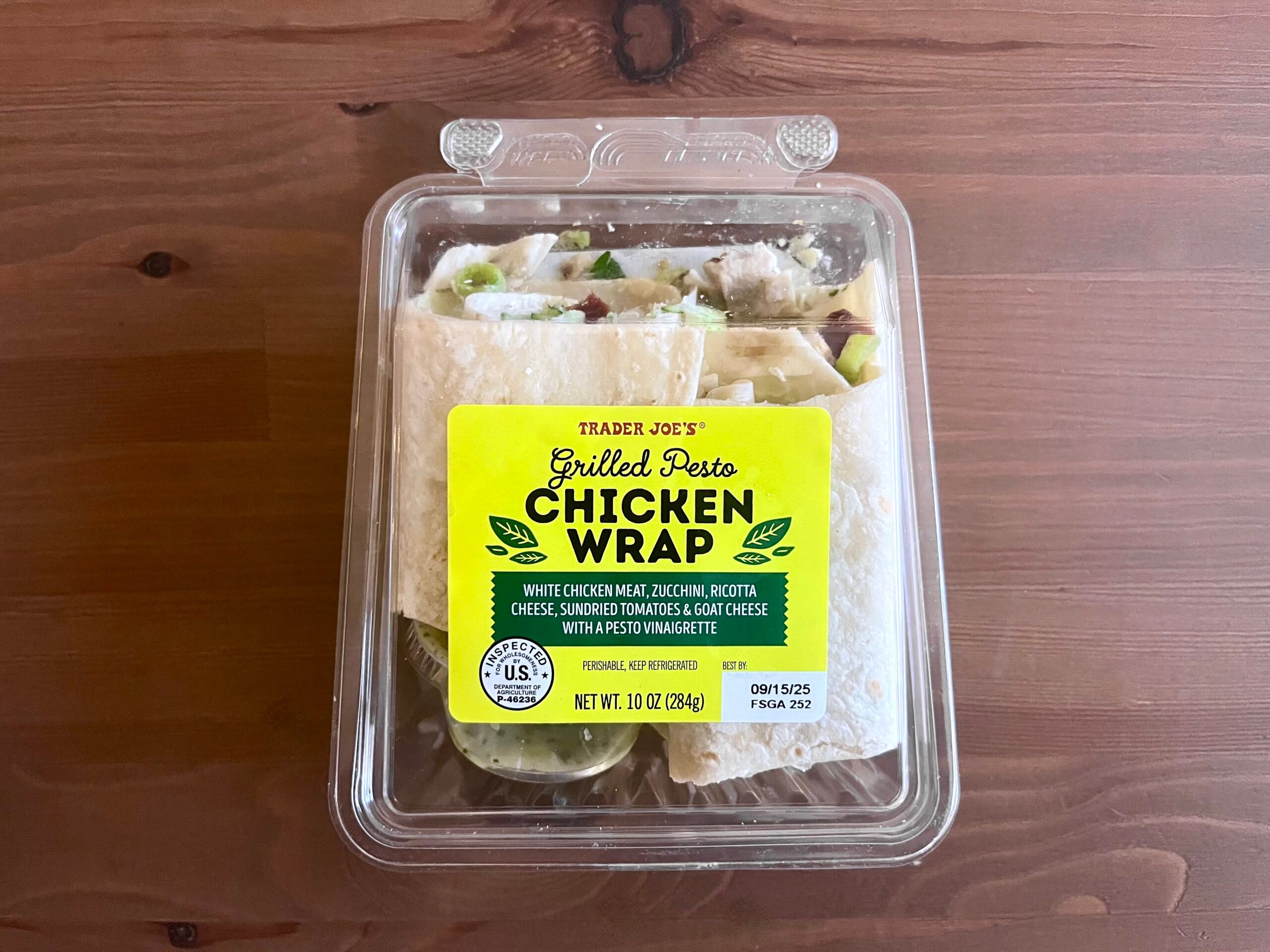 A clear plastic container of Trader Joe’s Grilled Pesto Chicken Wrap sits on a wooden table. The yellow label describes the wrap as containing chicken, zucchini, ricotta, sun-dried tomatoes, and oat cheese.