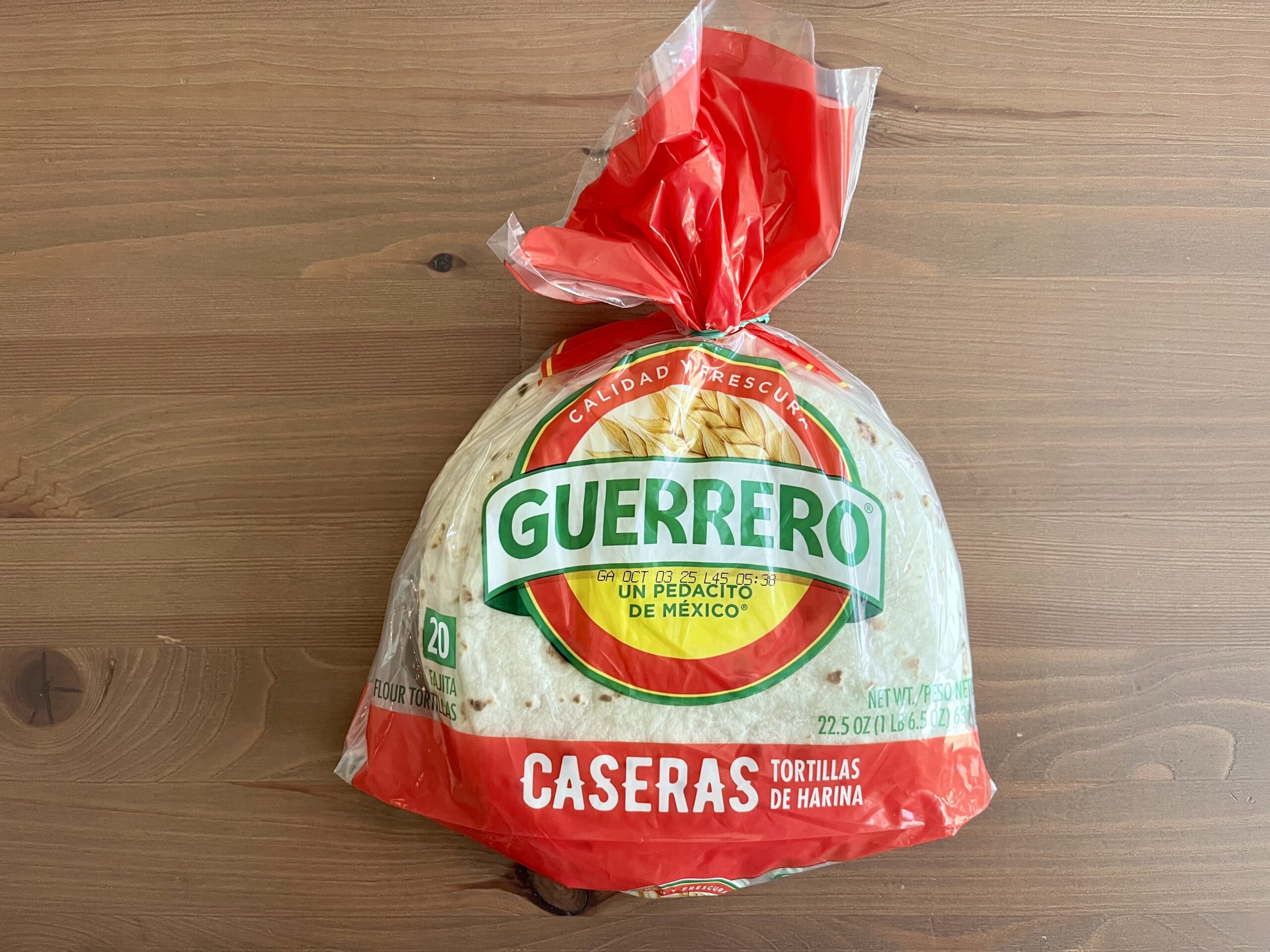 A package of Guerrero brand Caseras flour tortillas sits on a wooden surface. The package is clear with red and green labeling and contains 20 tortillas.
