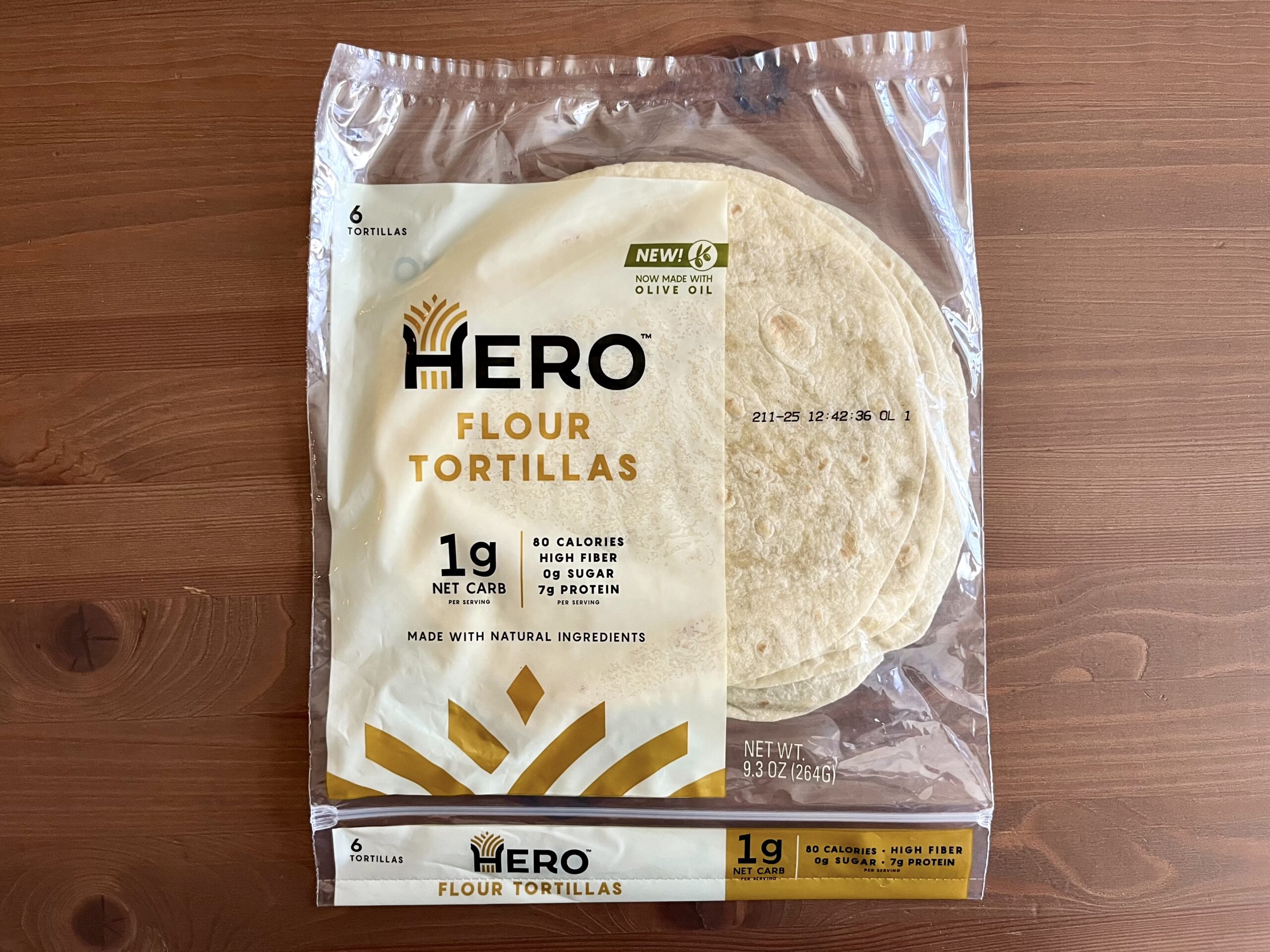 A package of Hero Flour Tortillas sits on a wooden surface. The label highlights 1g net carbs, high fiber, 0g sugar, and 7g protein per serving. Six tortillas are visible through the clear packaging.