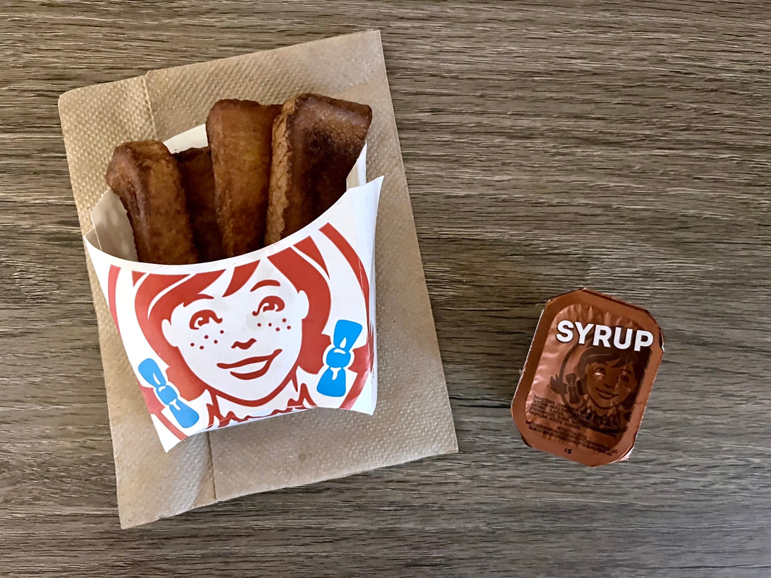 A container of French toast sticks in Wendy’s branded packaging sits on a napkin next to a sealed syrup cup on a wooden table.