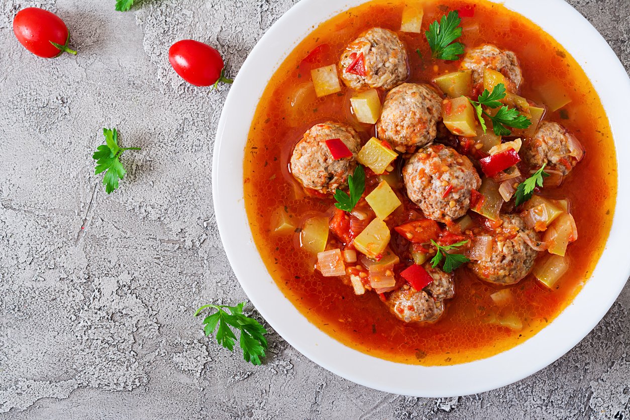 A white bowl filled with meatballs in a red tomato-based stew with diced vegetables, garnished with parsley. The bowl is on a gray textured surface, with tomatoes and parsley leaves nearby.