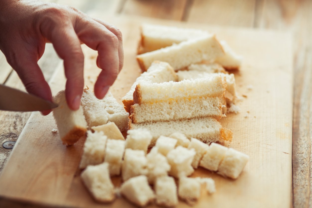 A hand slices white sandwich bread into cubes and strips on a wooden cutting board.