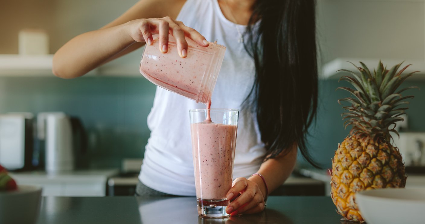 A woman pours a pink fruit smoothie from a blender into a glass in a kitchen, with a pineapple visible on the counter.