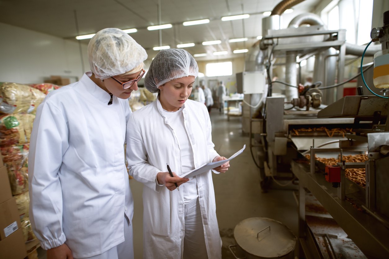 Two workers wearing white lab coats and hairnets review paperwork in a food processing factory, surrounded by machinery and snacks on conveyor belts. The environment is clean and industrial.