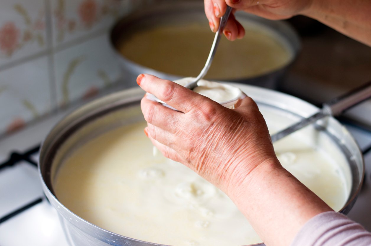 Close-up of hands using a spoon to transfer homemade yogurt or thick dairy mixture from a large pot into a small cup, with a stove and another pot in the background.