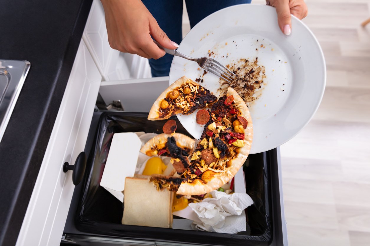 A person scrapes a burnt, partially eaten pizza from a plate into a kitchen trash can filled with other discarded food and paper waste.