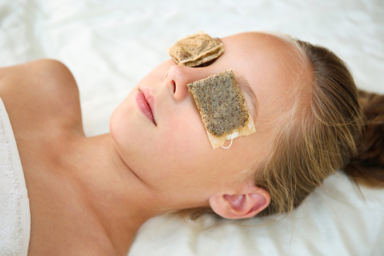 A young girl lies with her eyes closed, resting on a white surface, with used tea bags placed over her eyelids, suggesting a natural home remedy for eye care or relaxation.