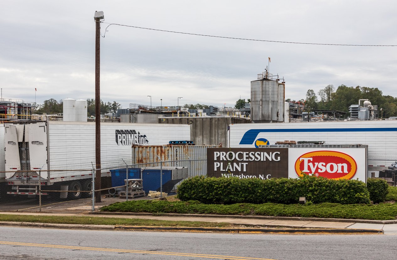 A Tyson Foods processing plant in WIlkesboro, NC, with a large sign out front. Several refrigeration trailers, including one with a “Prime” logo, are parked near the fenced facility on a cloudy day.