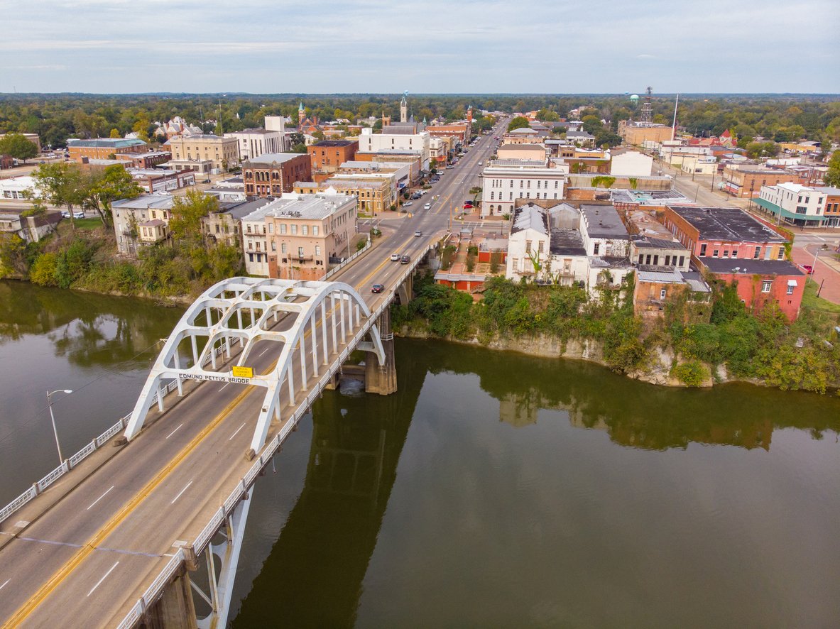 Aerial view of a bridge crossing over a river into a small town with historic buildings, trees, and cars on the road; the sky is partly cloudy.