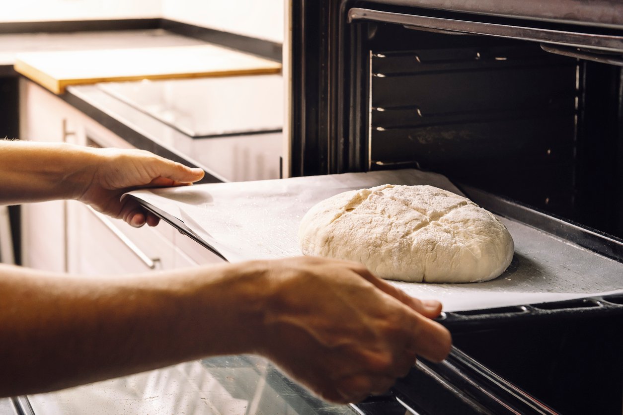 A person places a baking tray with a round, unbaked loaf of bread dough into an open oven in a kitchen.