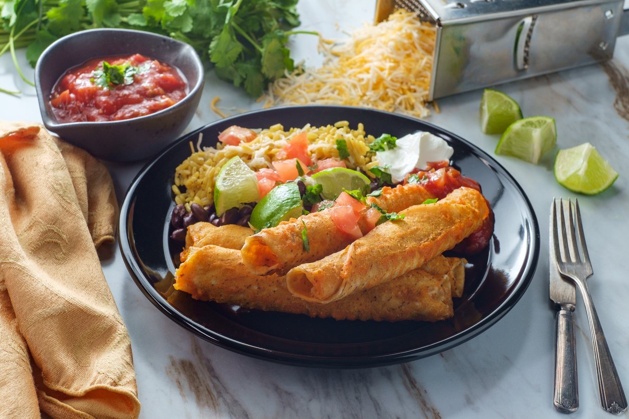 A black plate with three taquitos, Mexican rice, black beans, avocado, tomato, sour cream, and lime wedges. Salsa, shredded cheese, cilantro, a grater, and napkin are beside the plate on a marble surface.
