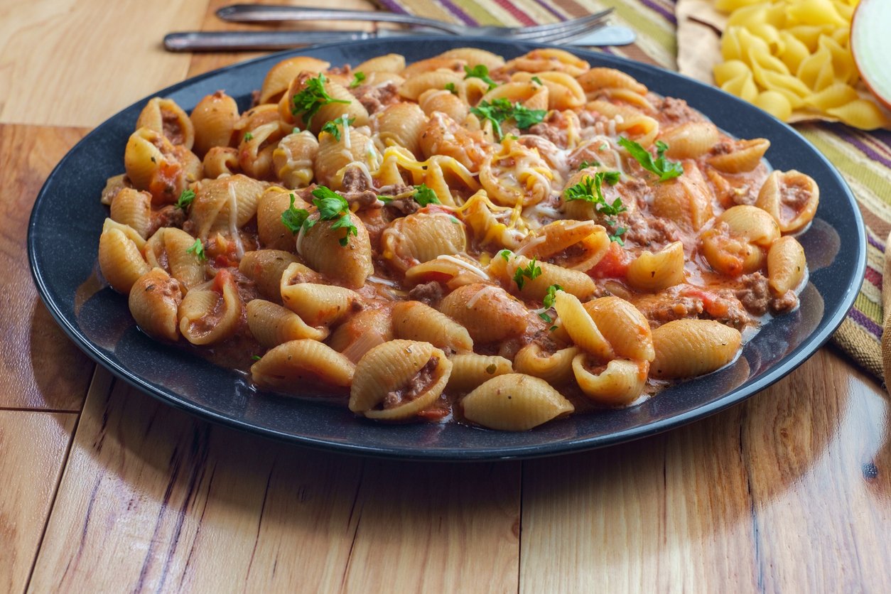 A black plate filled with shell pasta in a tomato meat sauce, garnished with chopped parsley, sits on a wooden table next to forks and a striped cloth napkin.