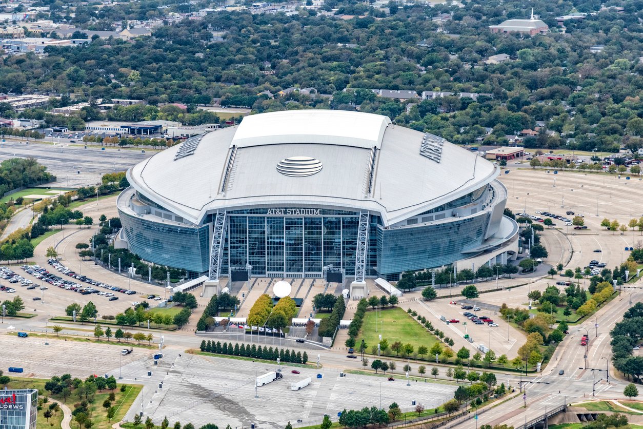 Aerial view of a large, modern stadium with a curved roof and glass façade, surrounded by parking lots, roads, and trees in an urban area.
