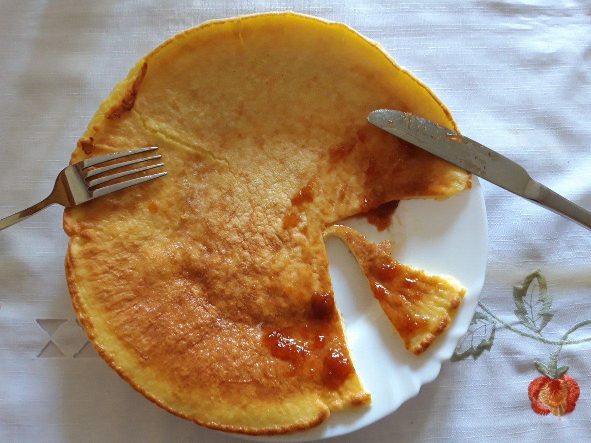 A large, thin pancake with jam sits on a white plate. A wedge has been cut and placed next to the main piece. A fork and knife rest on the plate, and the plate is on an embroidered white tablecloth.