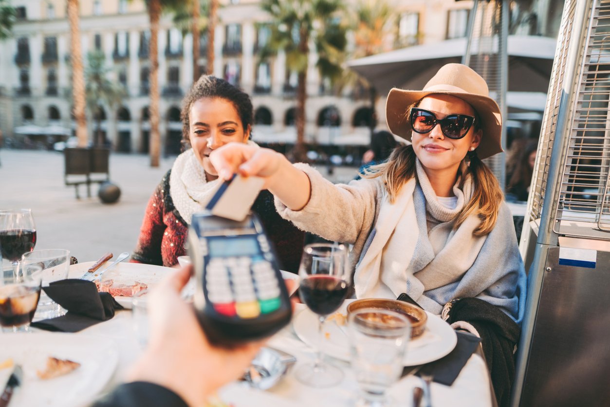 Two women sit at an outdoor restaurant table with food and wine. One woman, wearing sunglasses and a hat, smiles as she pays with a credit card using a wireless payment terminal. Palm trees and buildings are in the background.