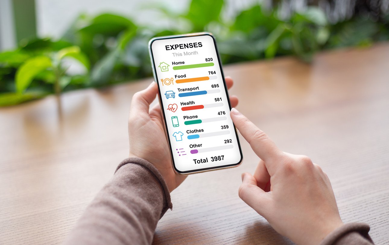 A person holds a smartphone displaying a colorful expenses breakdown for the month, listing categories like home, food, transport, health, phone, clothes, and other, with a total expense of 3987. Example of working toward financial independence.