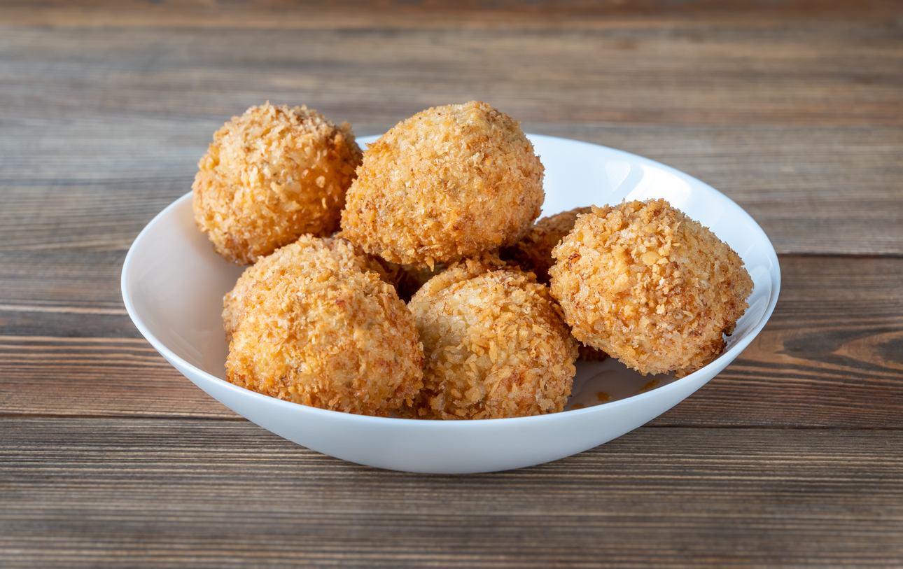 A white bowl filled with six golden-brown, crispy breaded balls sits on a wooden surface.