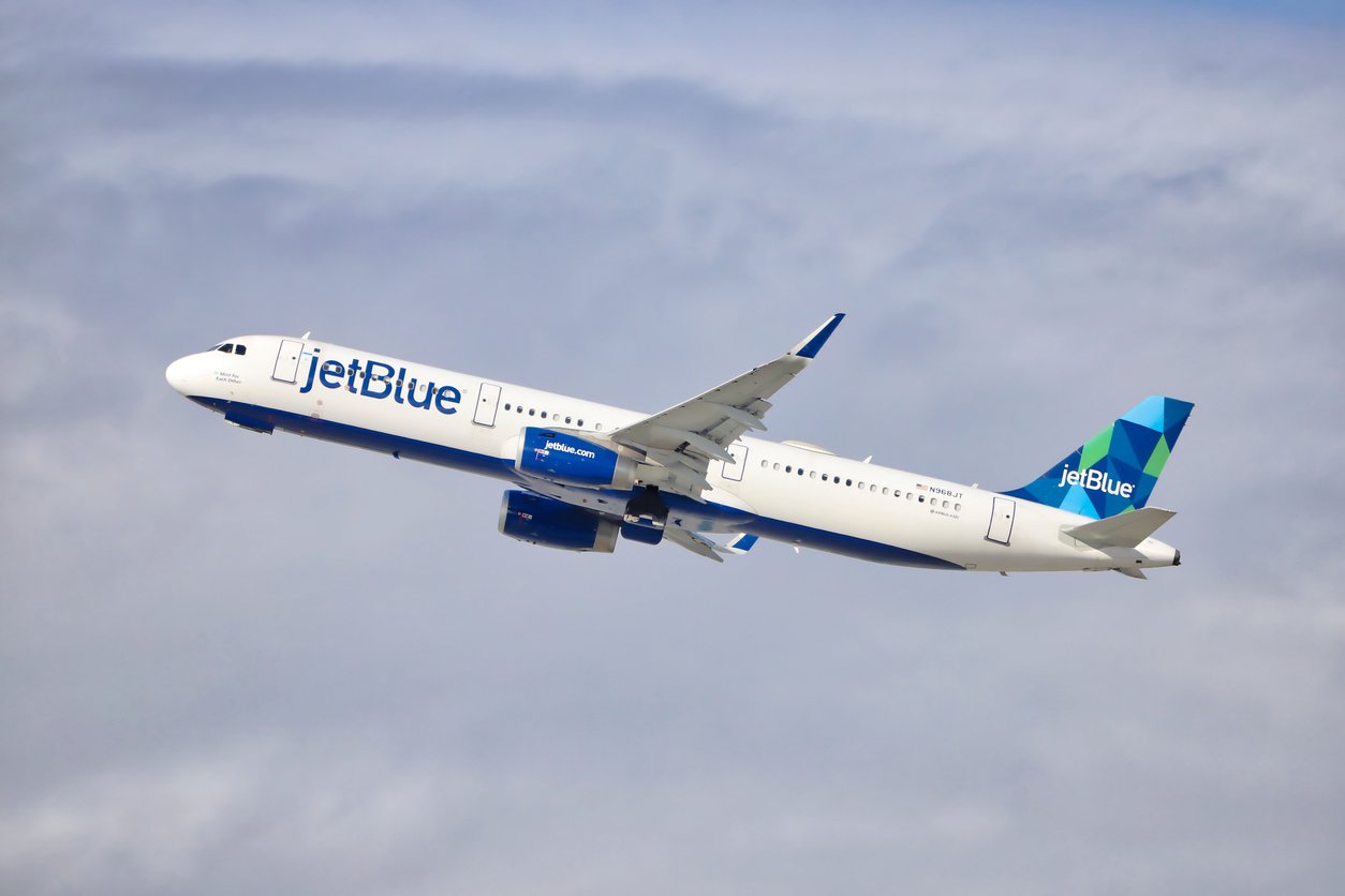 A JetBlue airplane with a blue and green tail fin is taking off, flying upward against a cloudy sky background.