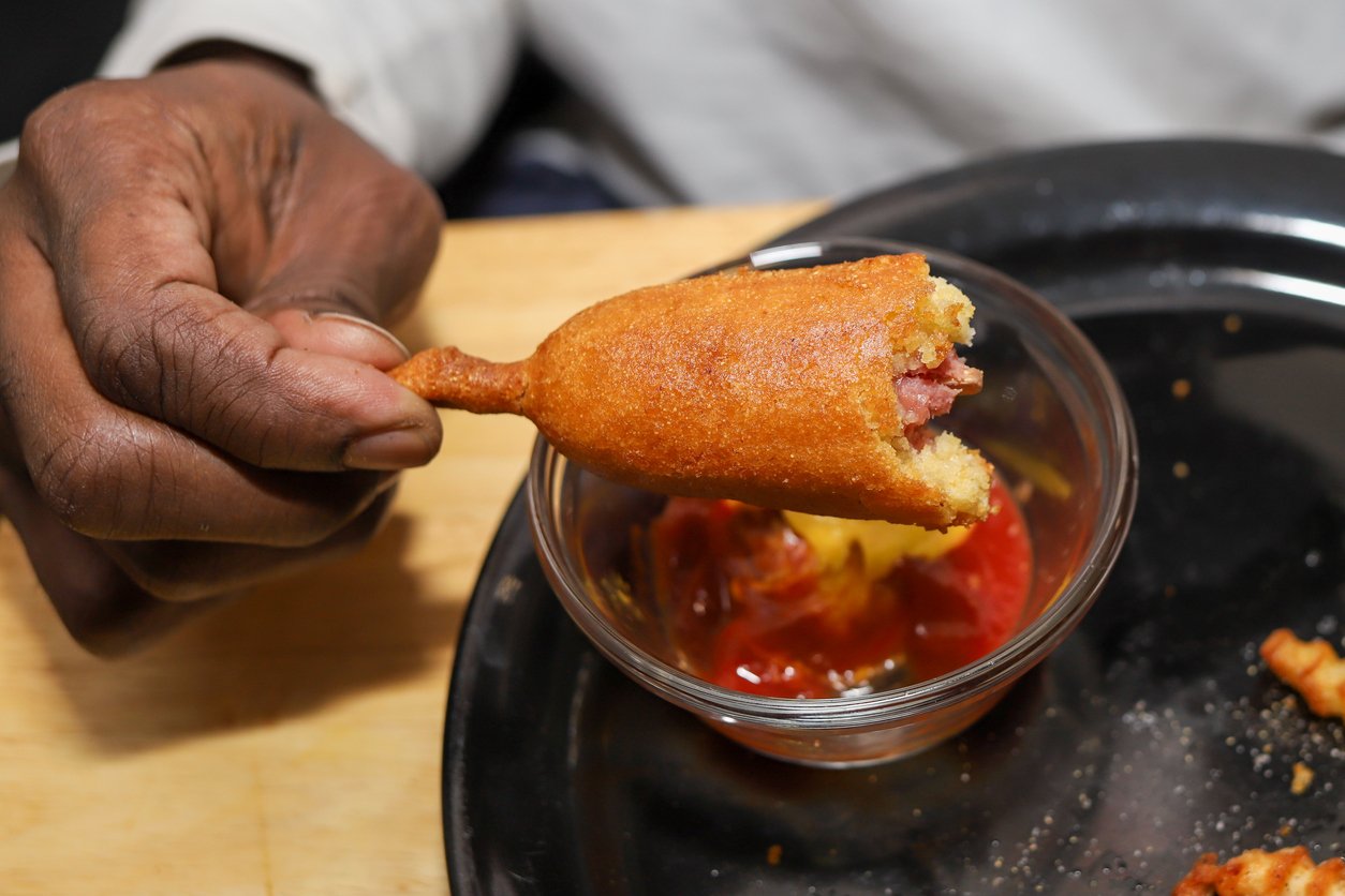 A hand holds a partially eaten corn dog above a small glass bowl of ketchup on a black plate, with crumbs and traces of ketchup scattered around—an image that brings to mind recent corn dog recall alerts.