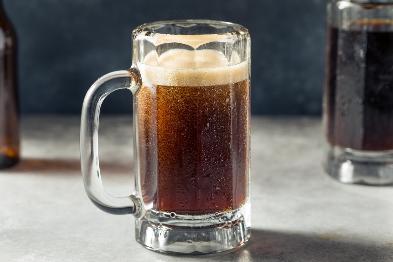 A glass mug filled with dark root beer sits on a gray surface, covered in condensation, with a foamy head at the top. Another mug and bottle are partially visible in the background.