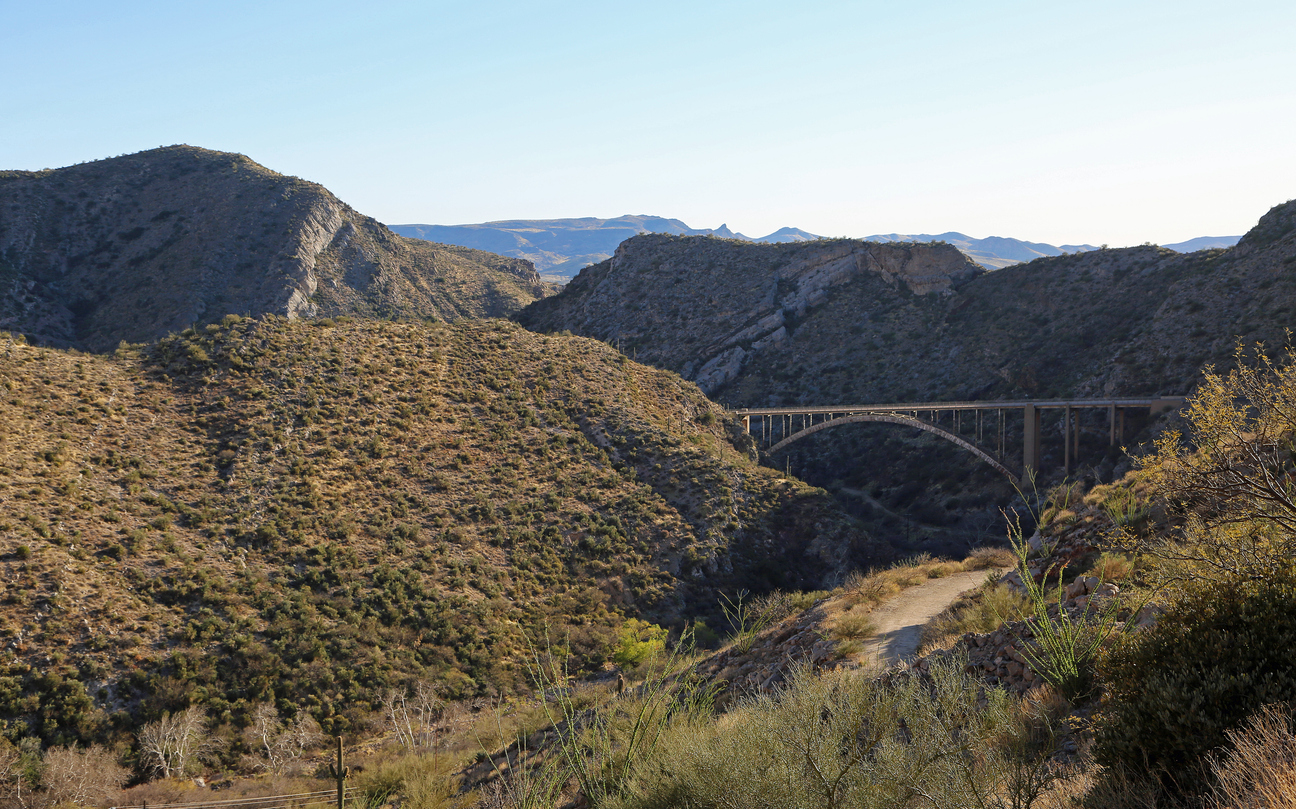 A steel arch bridge spans a deep, rugged canyon surrounded by dry, rocky hills and sparse desert vegetation under a clear blue sky.