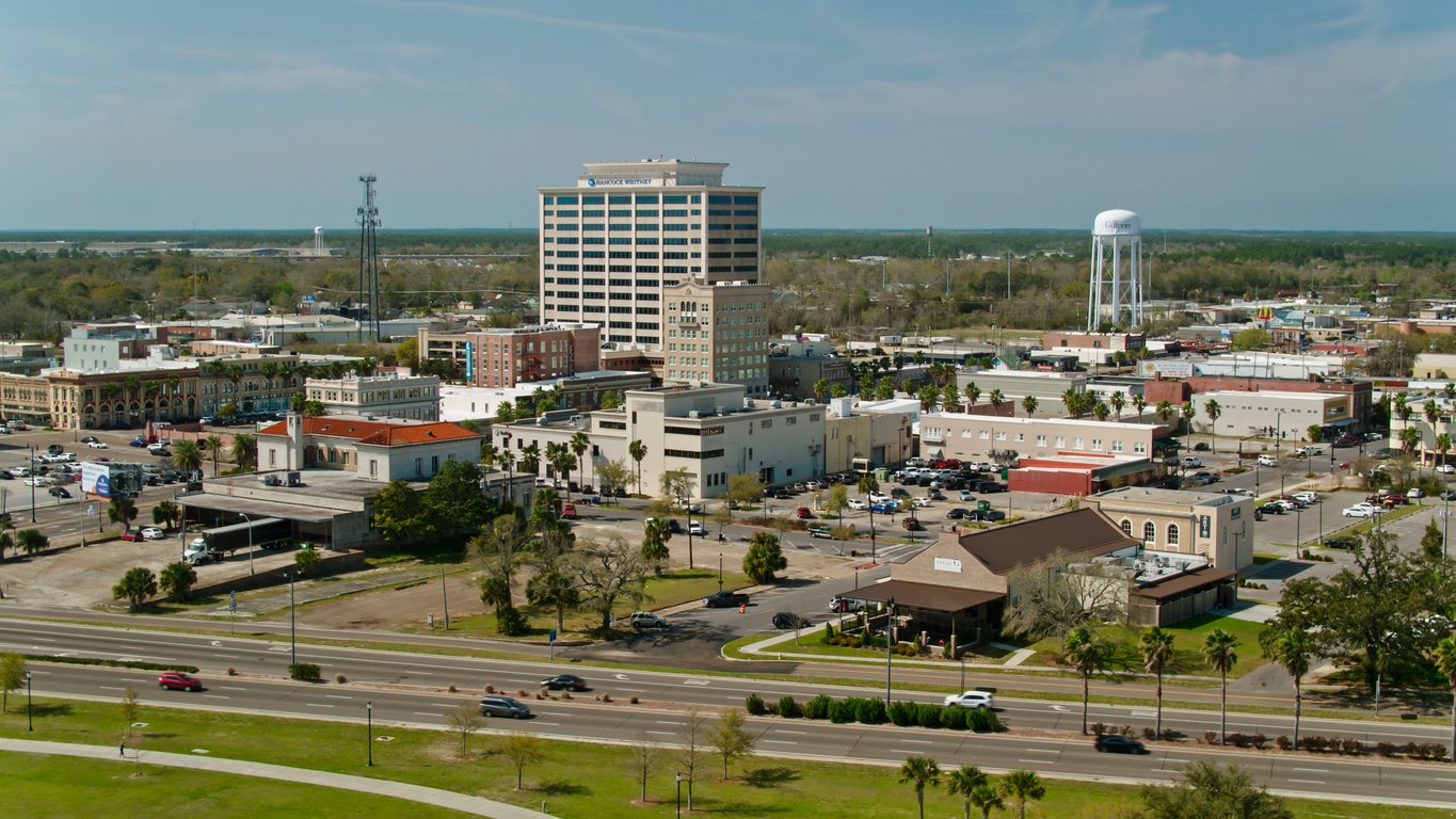 Aerial view of a mid-sized city with a mix of tall and low-rise buildings, roads, parked cars, greenery, and a large white water tower in the background under a partly cloudy sky.