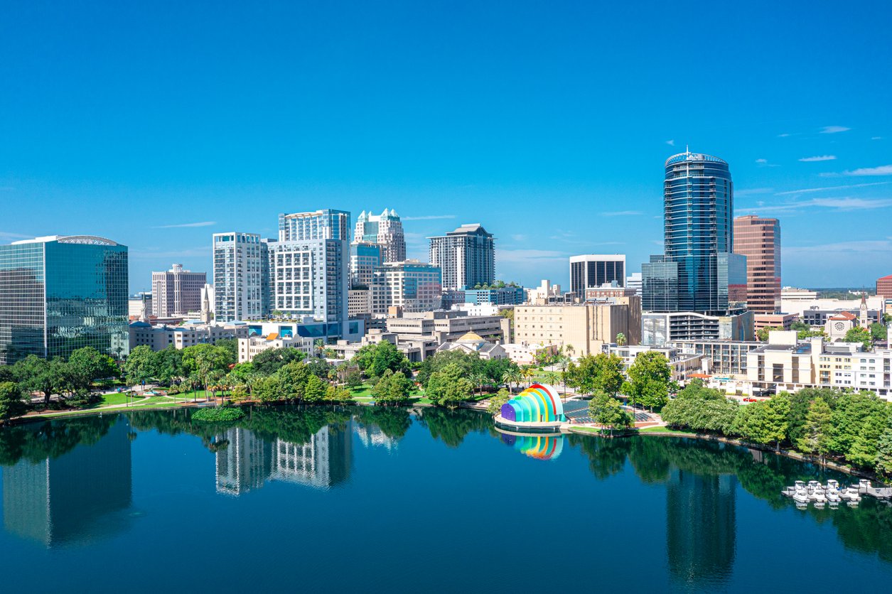 A clear blue sky over a modern city skyline with tall buildings, a reflective lake in the foreground, green trees, and a colorful rainbow structure by the water’s edge.