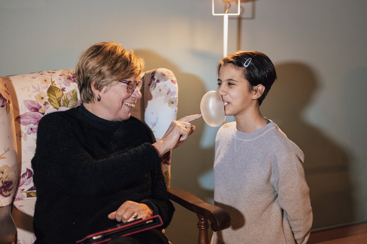 An older woman joyfully pops a bubblegum bubble blown by a smiling child with short dark hair, indoors on a floral armchair.