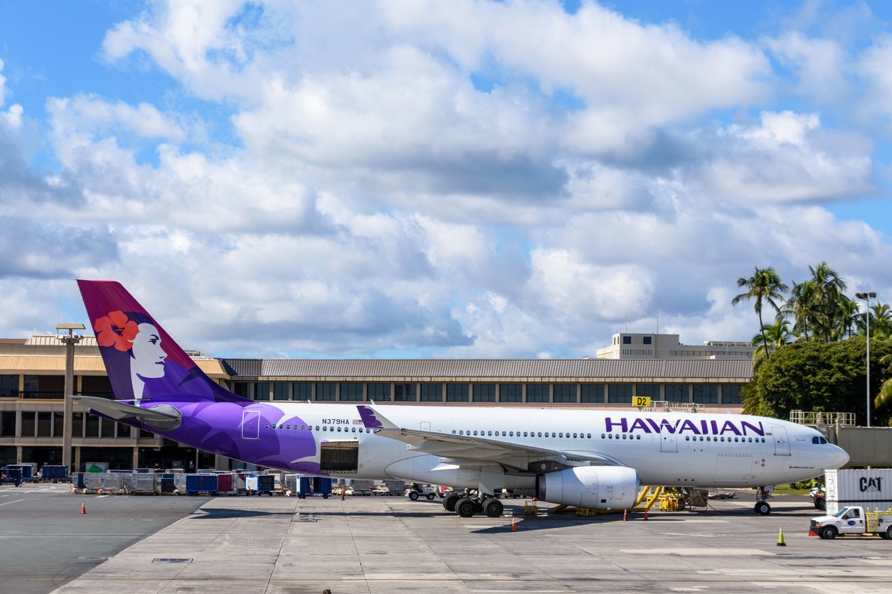 A Hawaiian Airlines plane with a purple tail and a flower design is parked on the airport tarmac in front of a terminal building, with blue sky and scattered clouds in the background.