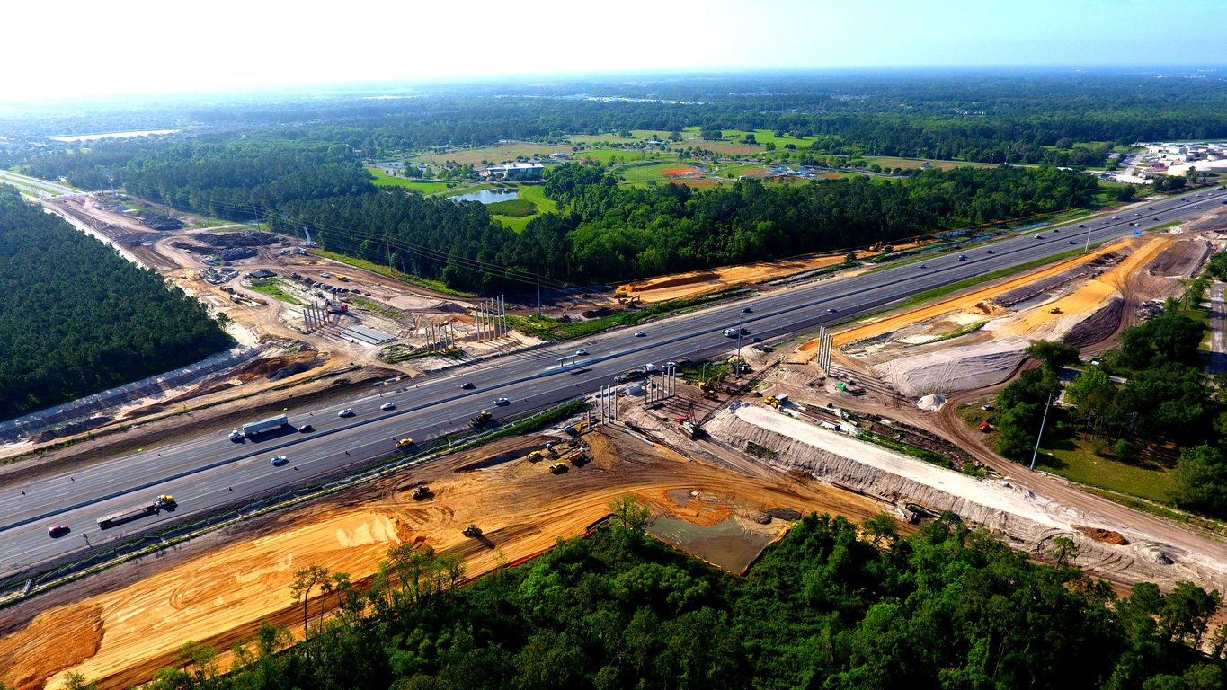 Aerial view of a highway under construction, with multiple lanes, construction vehicles, dirt mounds, and green forested areas on both sides under a clear sky.