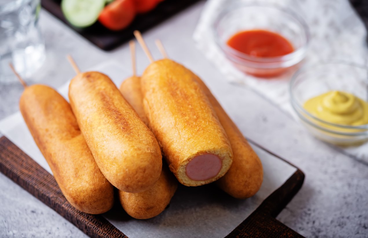 A pile of golden-brown corn dogs on a wooden board, with one corn dog showing the sausage inside. In the background, there are bowls of ketchup and mustard, and some sliced vegetables.