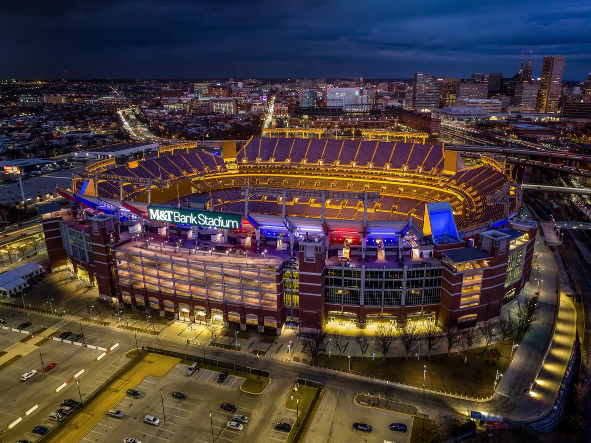 Aerial view of M&T Bank Stadium illuminated at night, surrounded by parking lots and city buildings, with the downtown skyline visible in the background.
