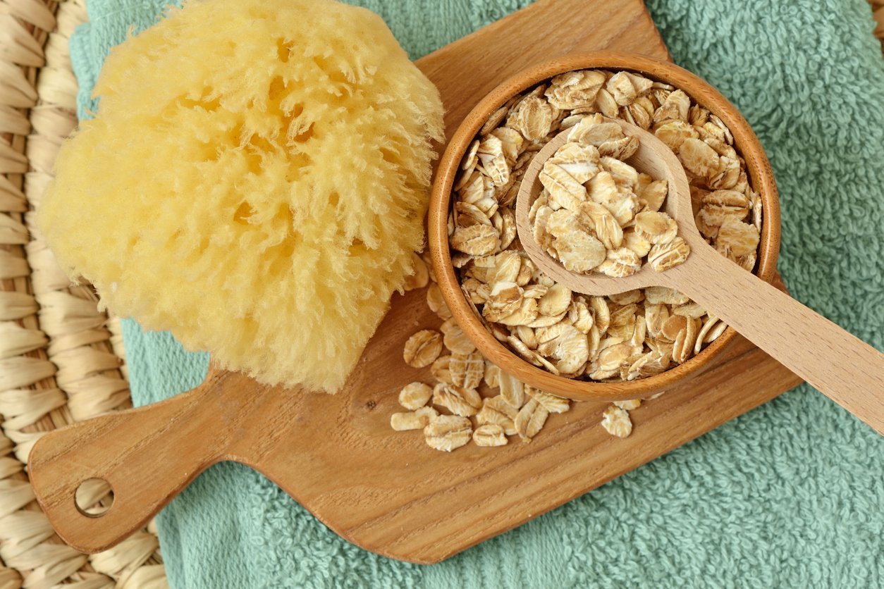 A wooden bowl filled with rolled oats and a small wooden spoon sits on a cutting board next to a yellow sponge, all placed on a folded green towel.