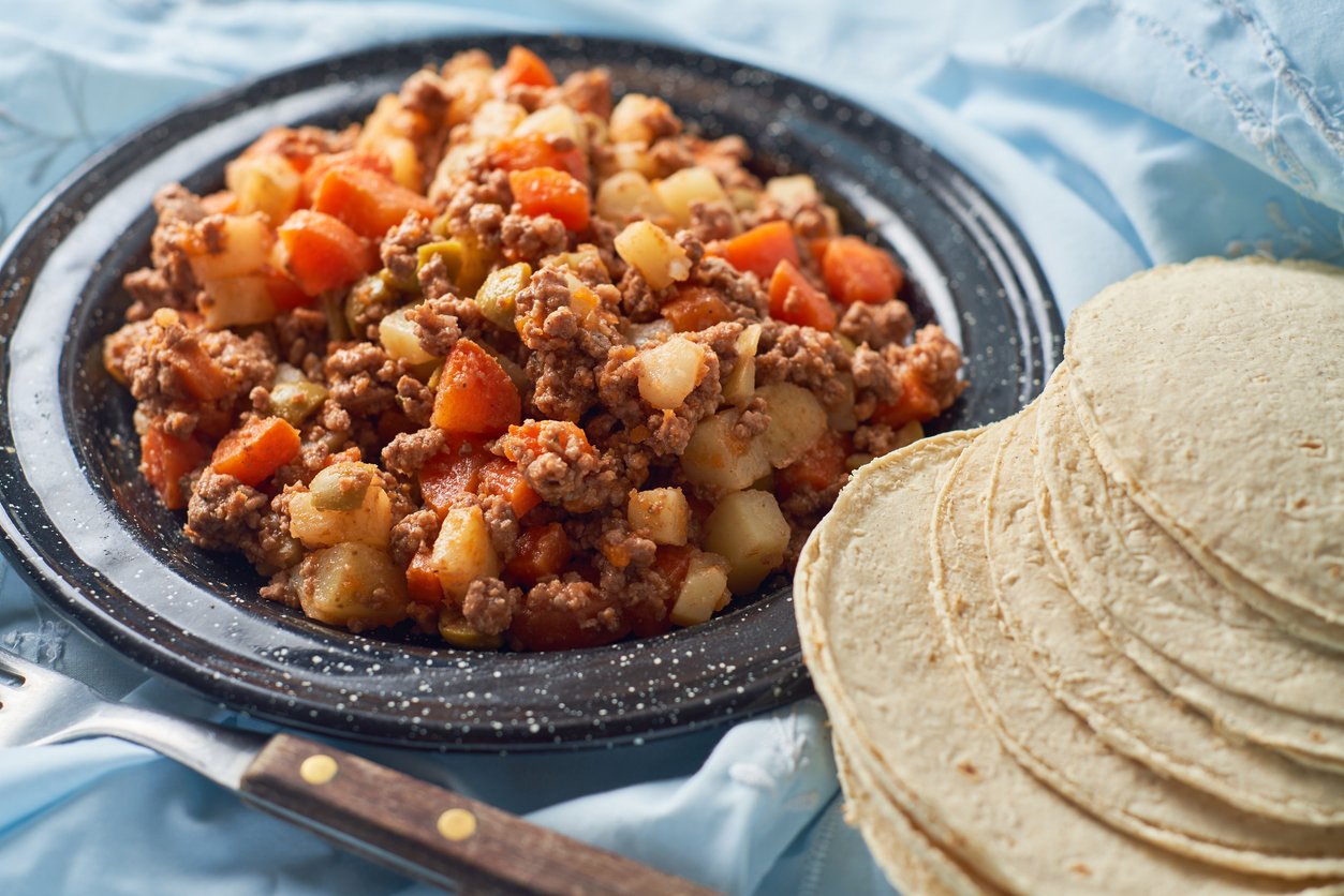 A plate of picadillo, a ground beef dish with diced potatoes, carrots, and vegetables, sits next to a stack of corn tortillas on a blue cloth, with a fork nearby.