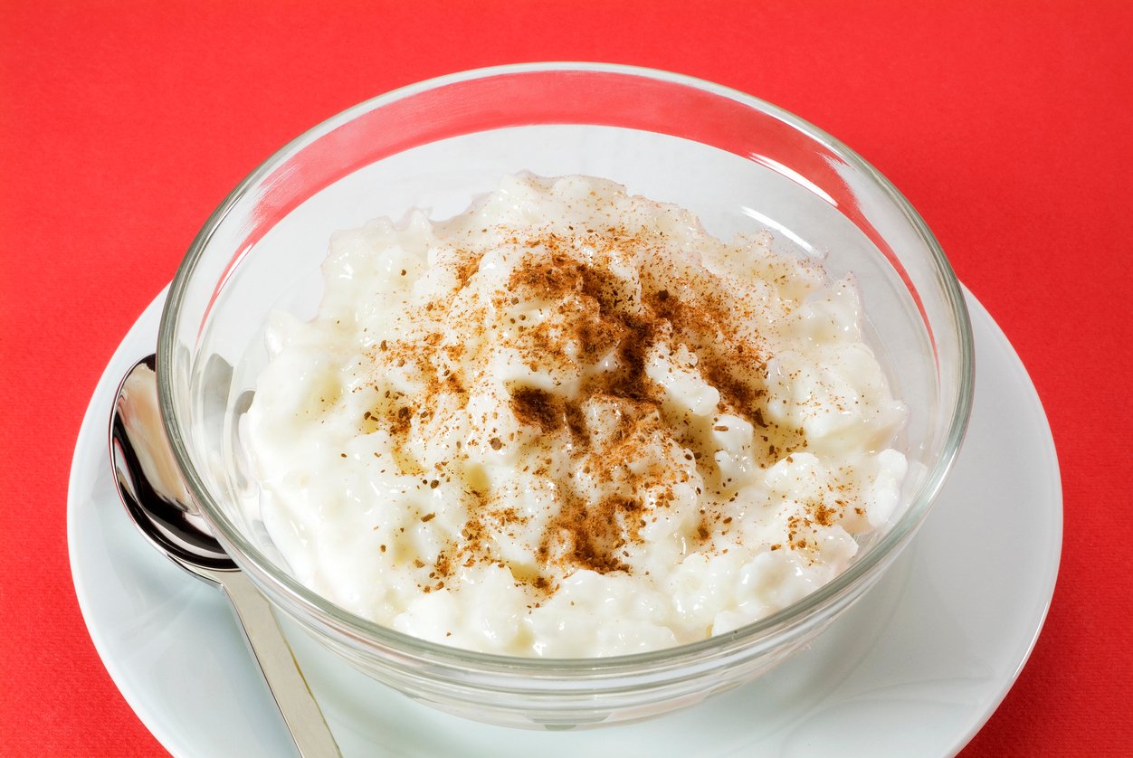 A glass bowl of rice pudding topped with ground cinnamon, placed on a white plate with a spoon, set against a bright red background.