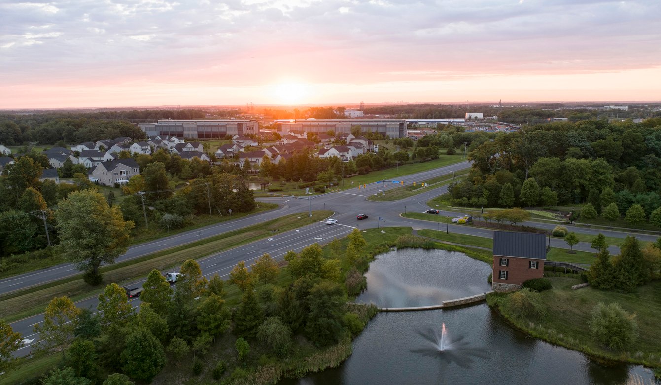 Aerial view of a suburban neighborhood at sunset with roads, houses, a small pond with a fountain, trees, and a distant building complex under a partly cloudy sky.
