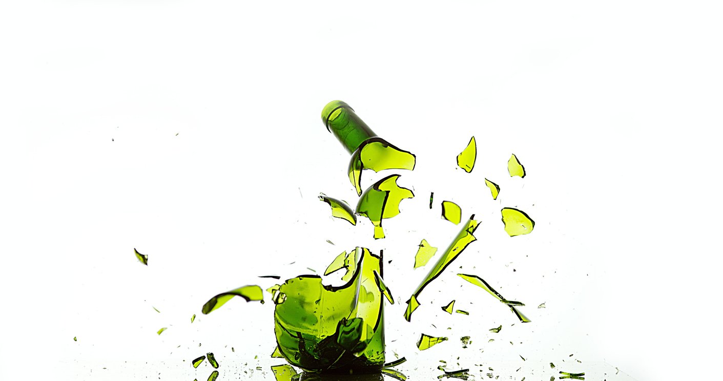 A green glass bottle shattering in mid-air, with fragments and pieces flying in different directions against a white background.