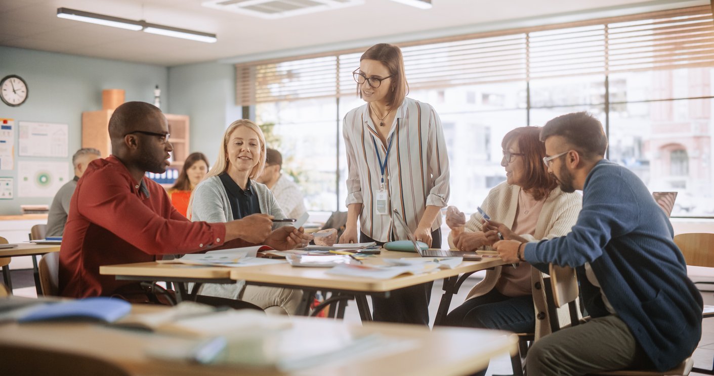 A group of adults sit around classroom tables, talking and smiling, while a woman stands and engages with them. The room is bright, with large windows, and teaching materials are spread across the tables.