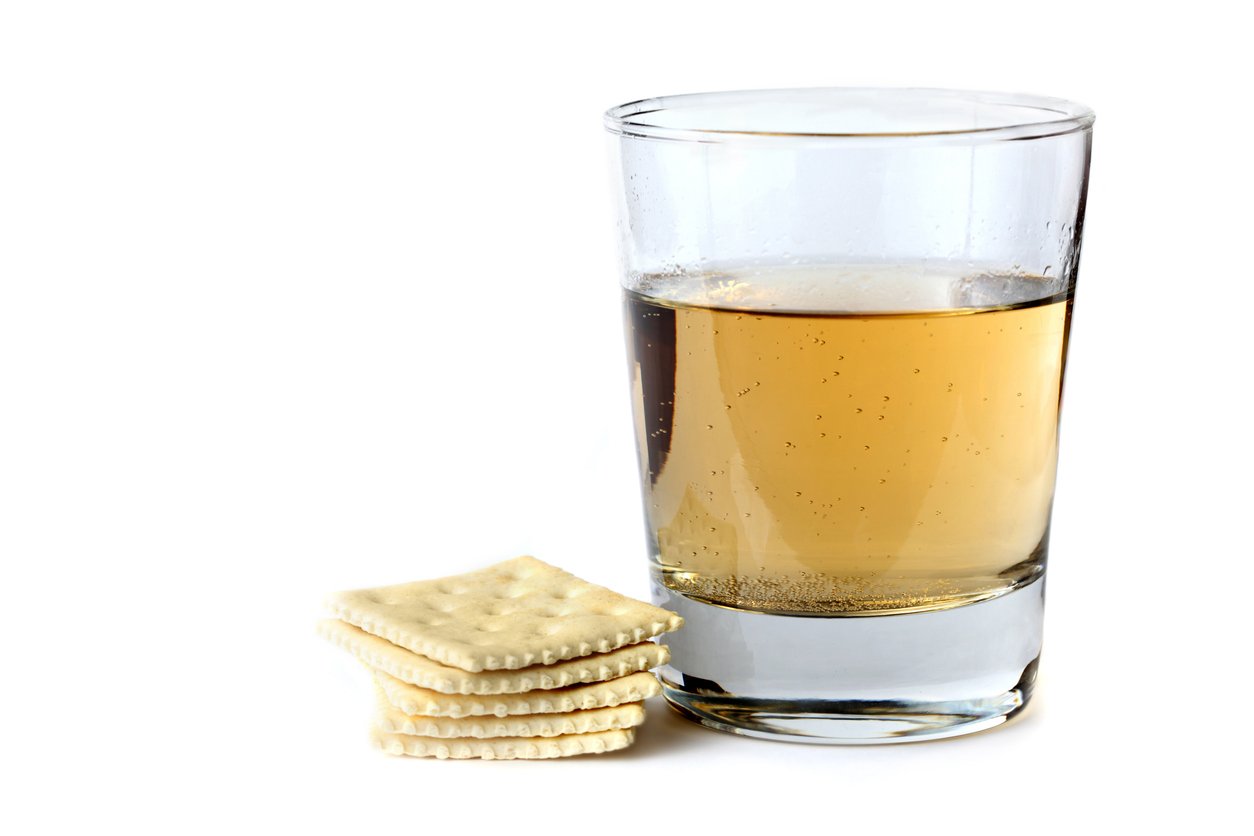 A clear glass of light brown liquid, possibly apple juice or tea, sits next to a small stack of square, salted crackers on a white background.