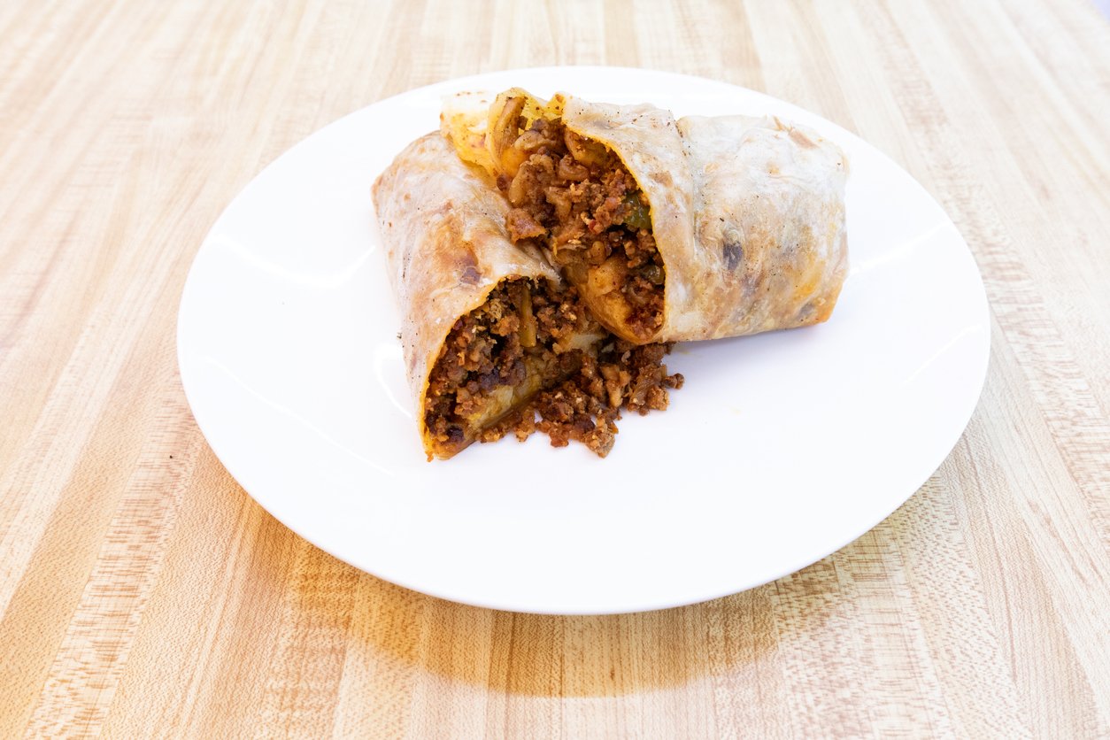 A white plate on a wooden table holds a burrito cut in half, revealing a filling of seasoned ground beef and spices.