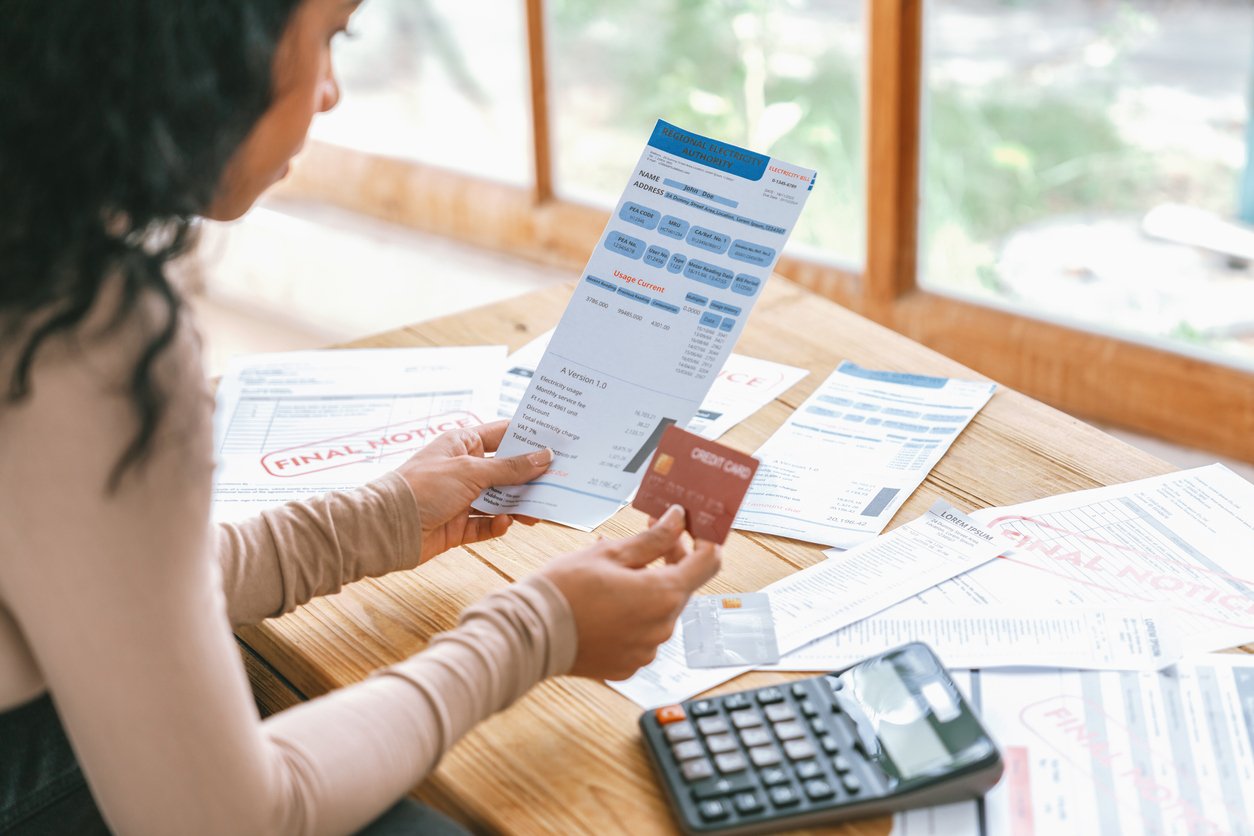 A person holding a credit card and a bill, surrounded by final notice documents on a wooden table, with a calculator nearby. Sunlight streams in through a window in the background.