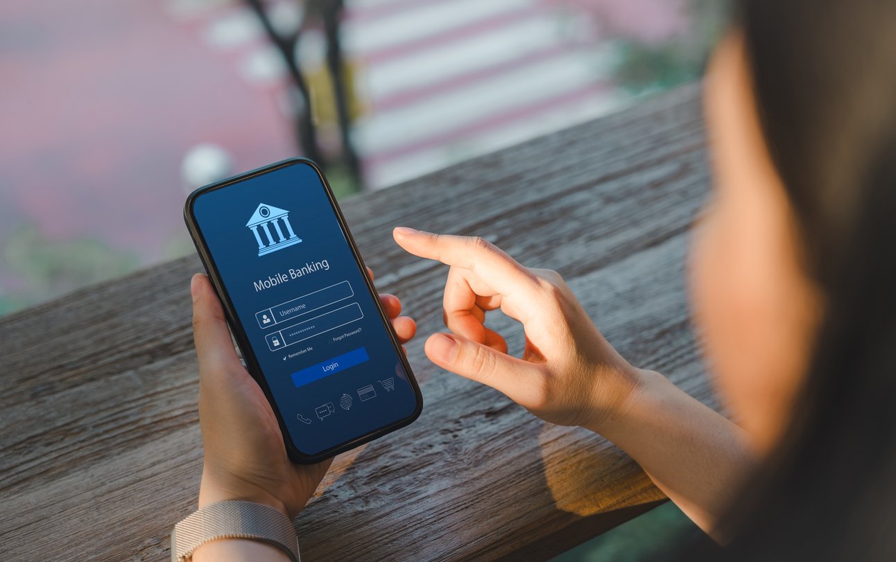 A person holds a smartphone and uses their finger to log in to a mobile banking app, with login fields for username and password visible on the screen. They are seated at a wooden counter near a window.