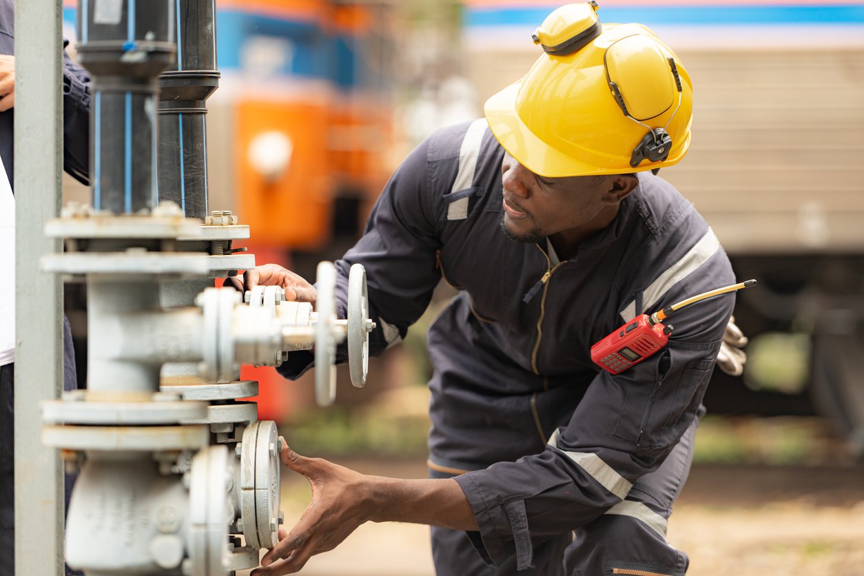 A worker wearing a yellow hard hat, safety goggles, and dark coveralls adjusts valves on industrial piping outdoors. He holds tools and has a walkie-talkie clipped to his arm. Trains are visible in the background.