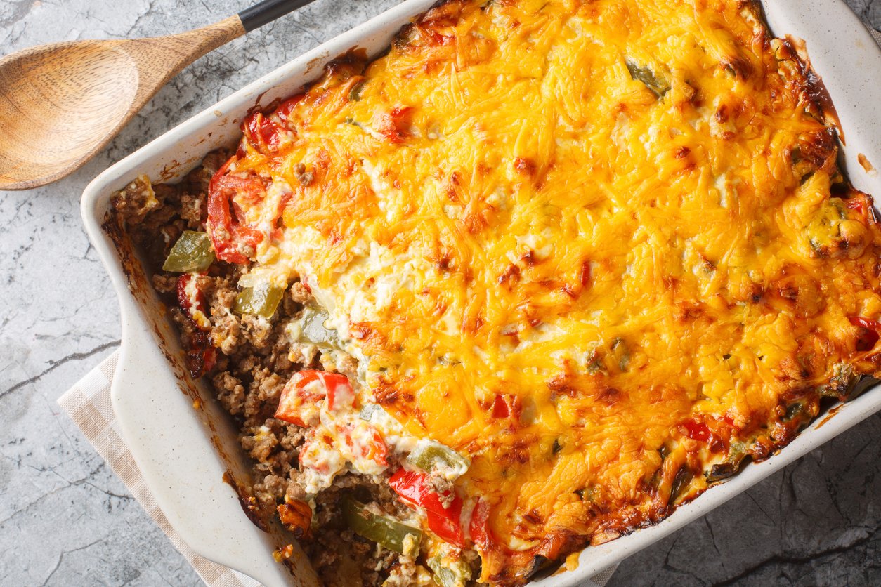 A casserole dish filled with a baked cheesy beef and vegetable casserole, partially served to reveal layers of ground meat, bell peppers, tomatoes, and melted cheese on top. A wooden spoon rests nearby on a gray surface.