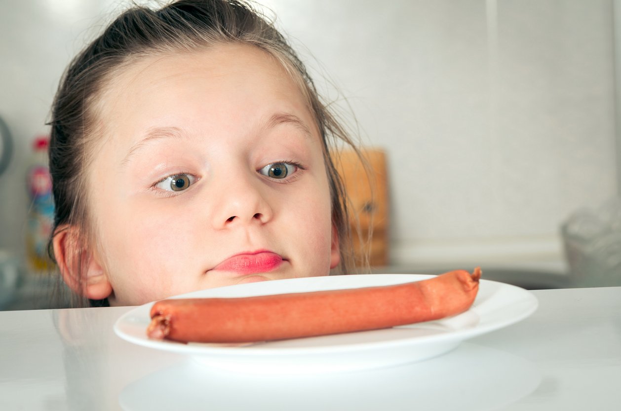 A young girl with wide eyes and a curious expression looks closely at a single sausage on a white plate in a kitchen setting.