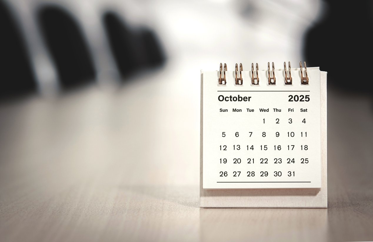 A small desk calendar displaying the month of October 2025 sits on a blurred office table, with empty chairs and a conference room in the background.