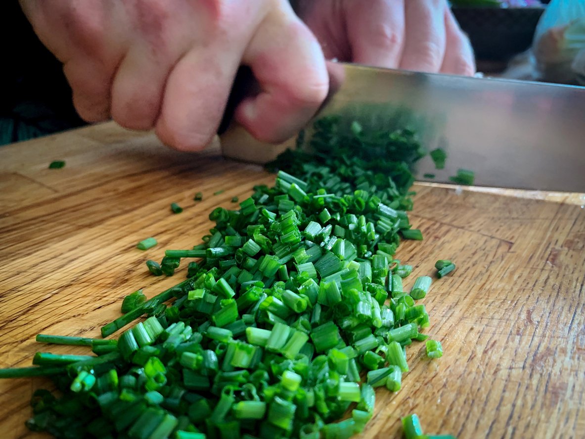 Close-up of hands using a knife to chop fresh green chives on a wooden cutting board. The chives are being cut into small pieces.