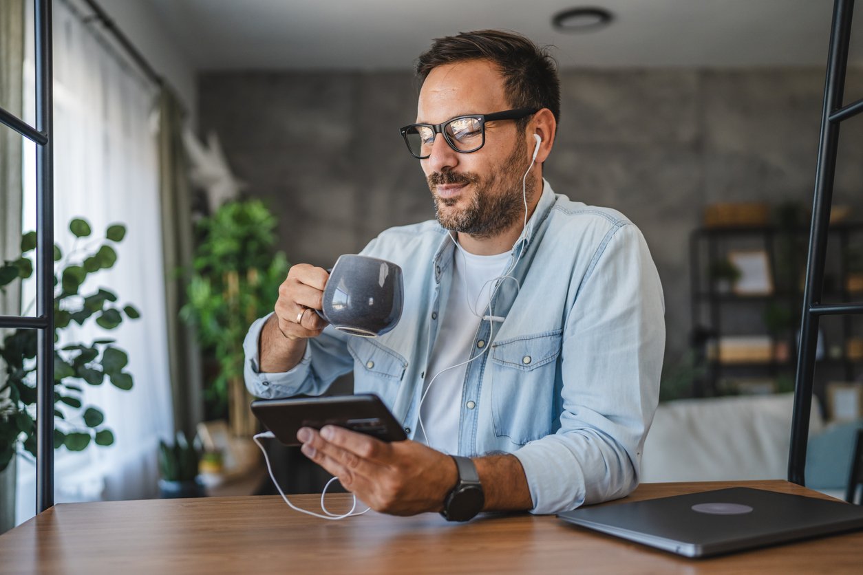 A man with glasses and earphones sits at a table, drinking from a mug and holding a tablet. A closed laptop is on the table, and the background shows a bright room with plants and shelves.